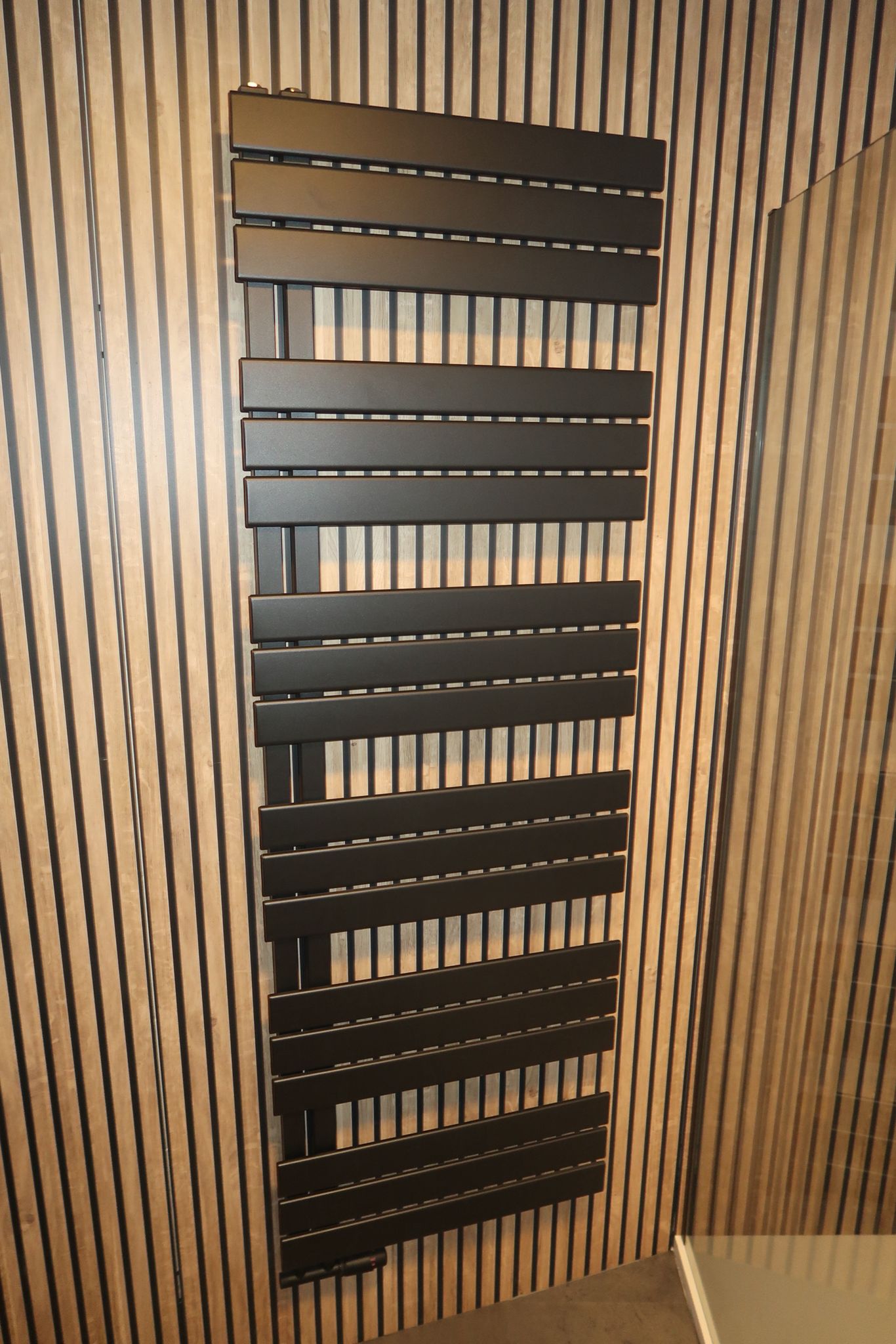Zwarte design handdoekradiator tegen een achtergrond van houtlook ribpanelen.